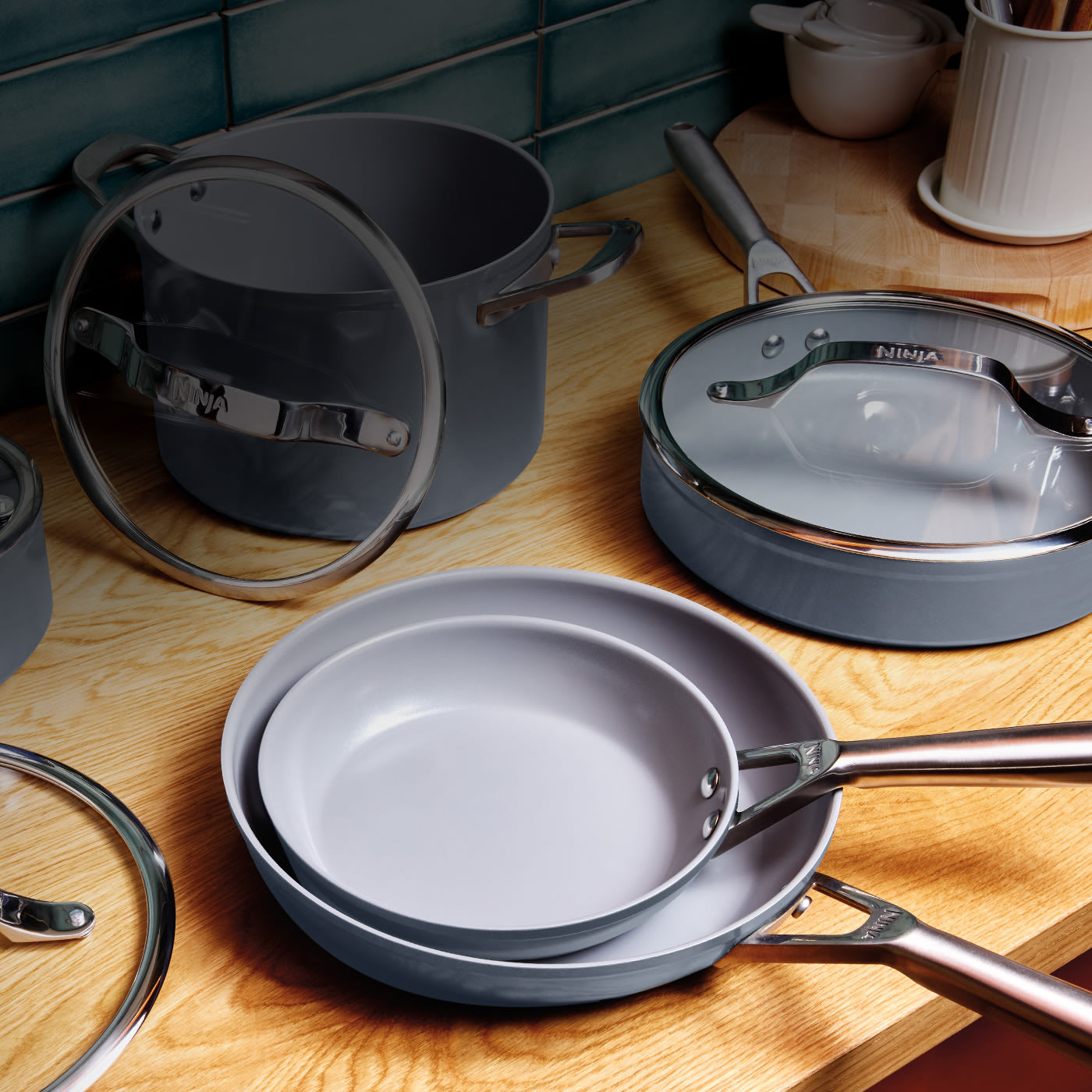 A collection of grey ceramic pro non-stick Ninja cookware, including saucepans, frying pans, and glass lids, displayed on a wooden kitchen worktop