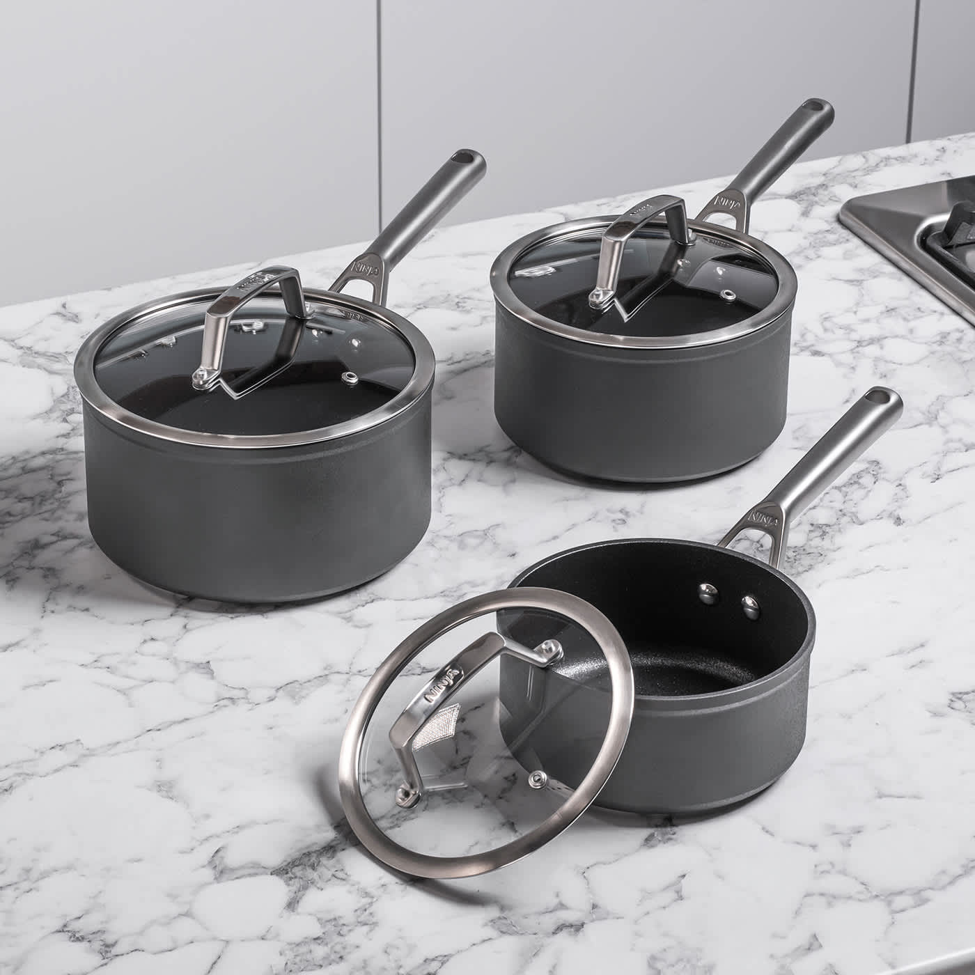 Image of Three Ninja saucepans with glass lids on a marble worktop.